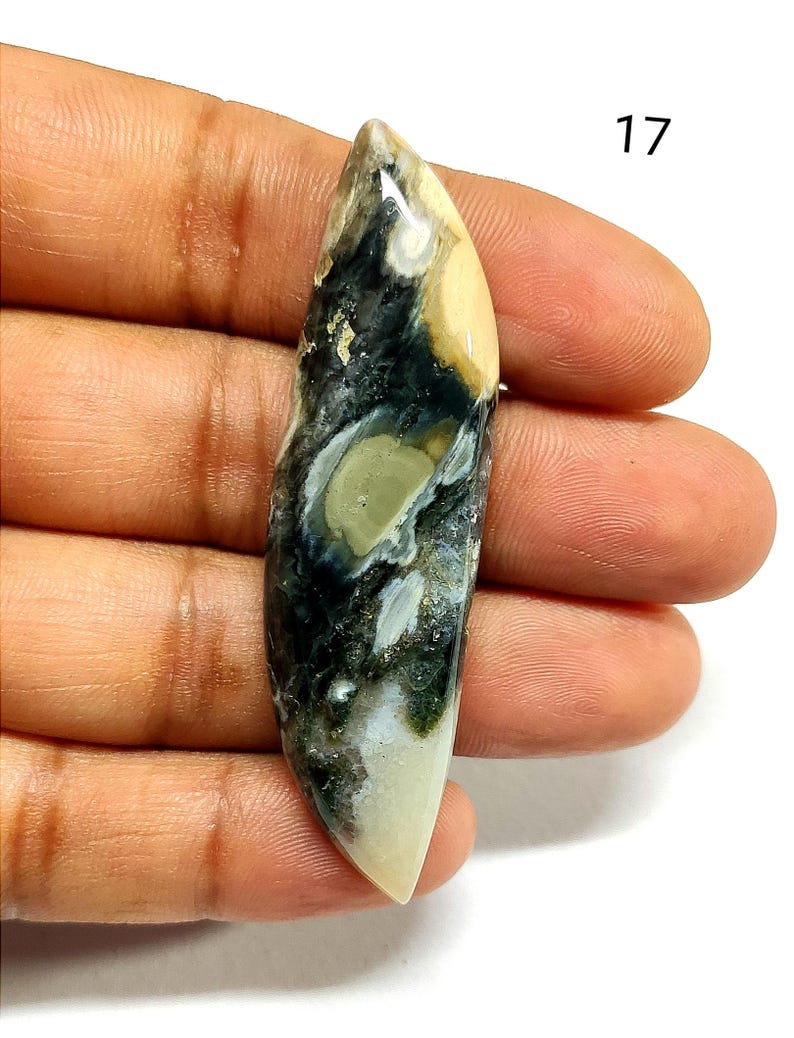 May include: An elongated, marquise-shaped polished gemstone cabochon featuring a marbled pattern of cream, dark charcoal, and sage green colors. The stone has a smooth, glossy surface with natural inclusions and the number 17 printed in black.