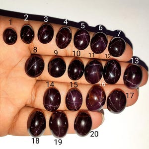 May include: A collection of twenty oval-shaped, dark red gemstones, each with a unique star-like pattern. The gemstones are displayed on a hand, showcasing their polished surfaces and varying sizes. The stones are numbered 1 through 20.