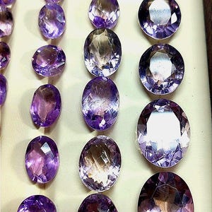 K&ouml;nnte beinhalten: Eine Auswahl an facettierten Amethyst-Edelsteinen in verschiedenen Formen und Gr&ouml;&szlig;en. Die Edelsteine reichen in der Farbe von hellem Lavendel bis zu tiefem Violett, mit ovalen und runden Schliffen. Die Steine werden auf einer hellen Oberfl&auml;che pr&auml;sentiert.