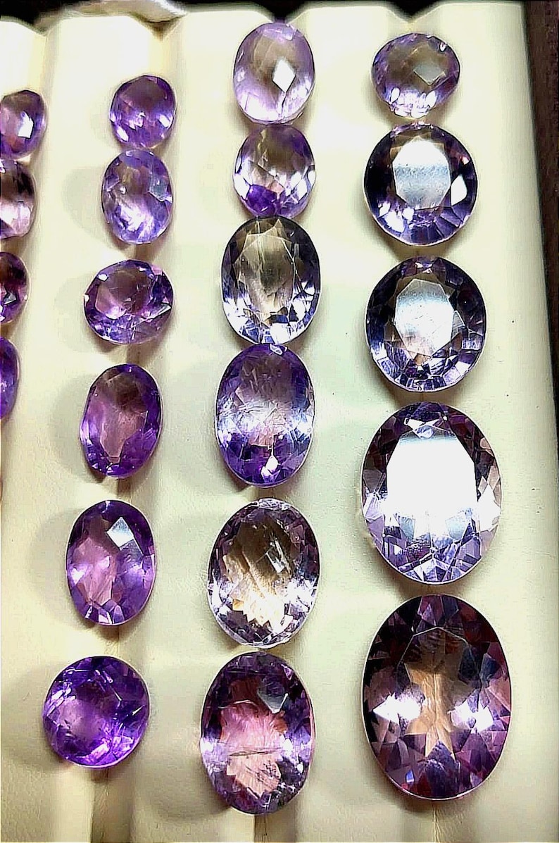 K&ouml;nnte beinhalten: Eine Auswahl an facettierten Amethyst-Edelsteinen in verschiedenen Formen und Gr&ouml;&szlig;en. Die Edelsteine reichen in der Farbe von hellem Lavendel bis zu tiefem Violett, mit ovalen und runden Schliffen. Die Steine werden auf einer hellen Oberfl&auml;che pr&auml;sentiert.