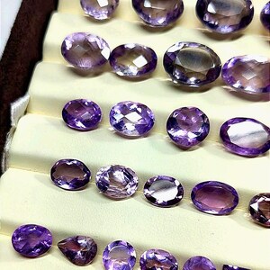 K&ouml;nnte beinhalten: Eine Auswahl an facettierten Amethyst-Edelsteinen in verschiedenen Formen, darunter oval, rund und quadratisch. Die Edelsteine werden auf einer mehrstufigen, cremefarbenen Oberfl&auml;che pr&auml;sentiert, die ihre tiefvioletten Farbt&ouml;ne und ihre innere Klarheit hervorhebt.
