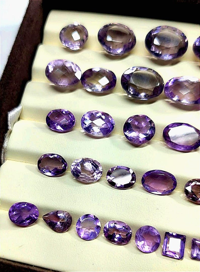 K&ouml;nnte beinhalten: Eine Auswahl an facettierten Amethyst-Edelsteinen in verschiedenen Formen, darunter oval, rund und quadratisch. Die Edelsteine werden auf einer mehrstufigen, cremefarbenen Oberfl&auml;che pr&auml;sentiert, die ihre tiefvioletten Farbt&ouml;ne und ihre innere Klarheit hervorhebt.