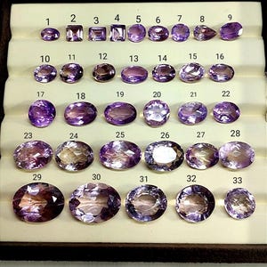 K&ouml;nnte beinhalten: Eine Sammlung von 33 Amethyst-Edelsteinen in verschiedenen Formen und Gr&ouml;&szlig;en, pr&auml;sentiert in einer braunen Schale. Die Edelsteine reichen von hell- bis tiefviolett, einige zeigen innere Muster. Jeder Stein ist zur Identifizierung nummeriert.