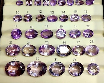 May include: A collection of 33 amethyst gemstones in various shapes and sizes, displayed in a brown tray. The gemstones range in color from light to deep purple, with some showing internal patterns. Each stone is numbered for identification.