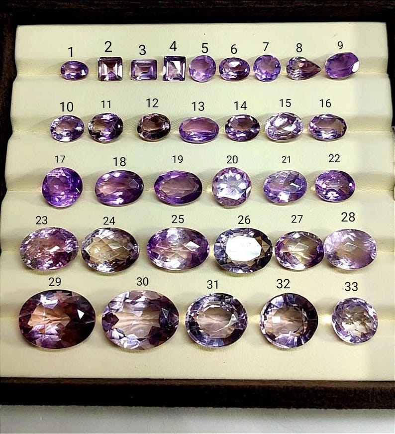 K&ouml;nnte beinhalten: Eine Sammlung von 33 Amethyst-Edelsteinen in verschiedenen Formen und Gr&ouml;&szlig;en, pr&auml;sentiert in einer braunen Schale. Die Edelsteine reichen von hell- bis tiefviolett, einige zeigen innere Muster. Jeder Stein ist zur Identifizierung nummeriert.