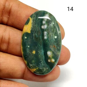 May include: An oval-shaped polished gemstone cabochon featuring a deep forest green base color with prominent mustard yellow patches and small white circular inclusions. The stone has a smooth, reflective surface and is marked with the number 14.