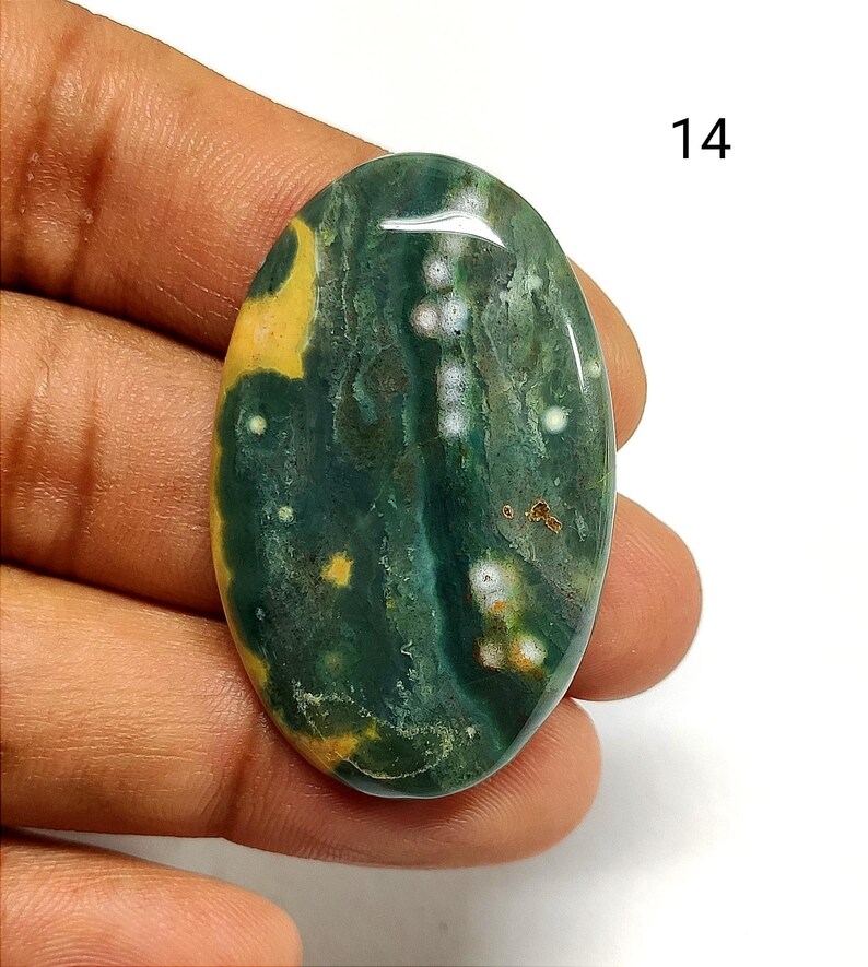 May include: An oval-shaped polished gemstone cabochon featuring a deep forest green base color with prominent mustard yellow patches and small white circular inclusions. The stone has a smooth, reflective surface and is marked with the number 14.