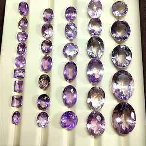 K&ouml;nnte beinhalten: Eine Sammlung von Amethyst-Edelsteinen in verschiedenen Formen und Gr&ouml;&szlig;en, darunter ovale, runde und rechteckige Schliffe. Die Edelsteine werden in einer braunen, samtgef&uuml;tterten Schale pr&auml;sentiert, die ihre tiefvioletten Farbt&ouml;ne und ihre innere Klarheit hervorhebt.