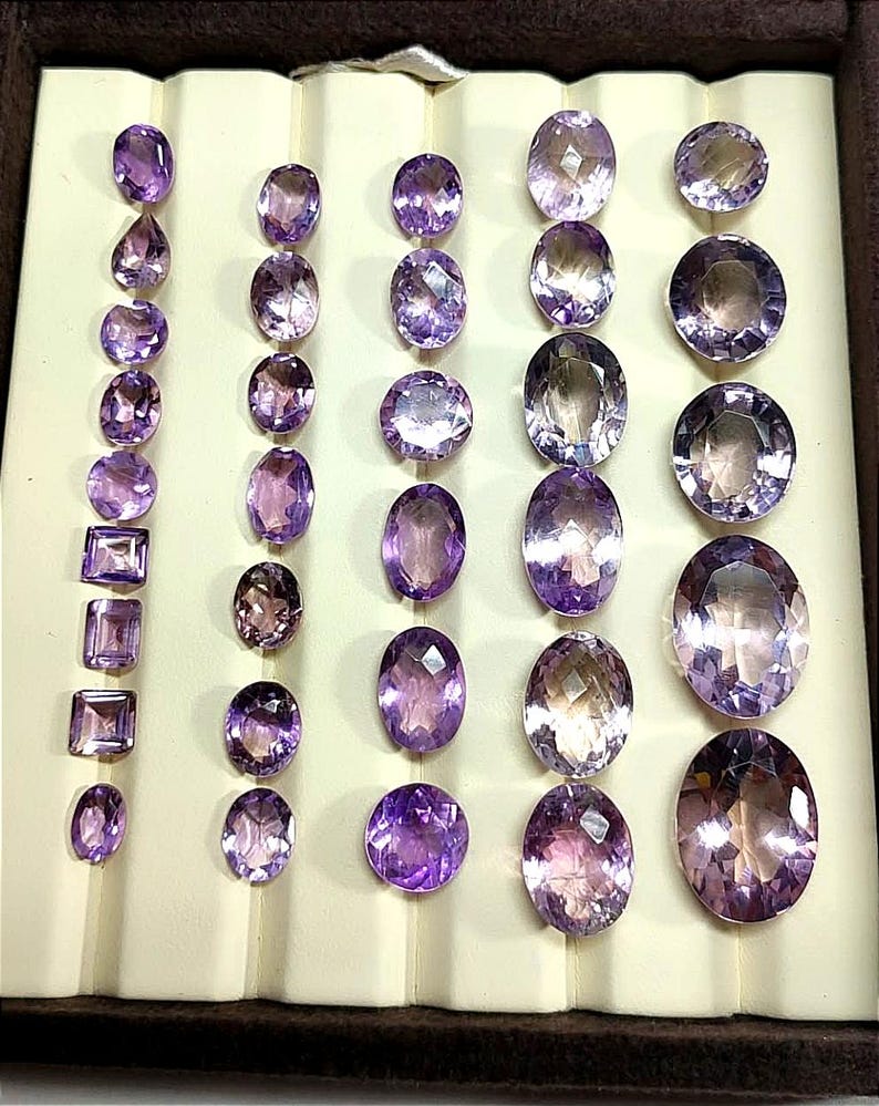 K&ouml;nnte beinhalten: Eine Sammlung von Amethyst-Edelsteinen in verschiedenen Formen und Gr&ouml;&szlig;en, darunter ovale, runde und rechteckige Schliffe. Die Edelsteine werden in einer braunen, samtgef&uuml;tterten Schale pr&auml;sentiert, die ihre tiefvioletten Farbt&ouml;ne und ihre innere Klarheit hervorhebt.