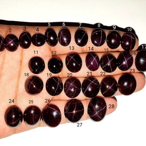 Könnte beinhalten: Eine Sammlung von achtundzwanzig dunkelroten, ovalen Cabochon-Edelsteinen, die in Reihen angeordnet sind und jeweils mit einer kleinen schwarzen Zahl von 1 bis 28 versehen sind. Die Steine zeigen ein vierstrahliges Sternmuster.