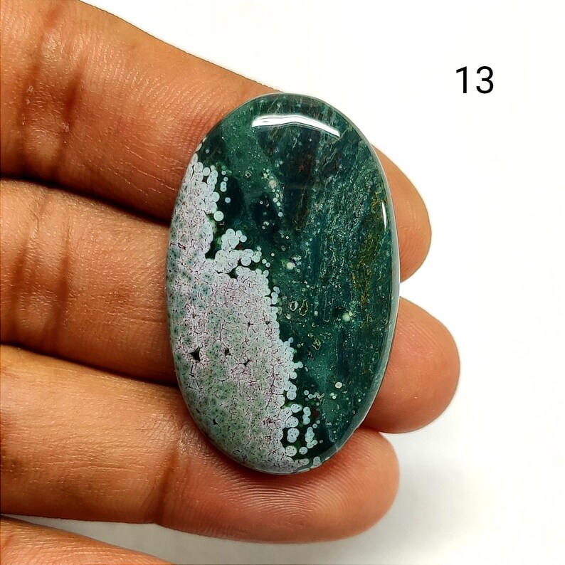 May include: An oval-shaped polished gemstone featuring a deep forest green base with a prominent white and light purple speckled pattern on the left side. The stone has a smooth, reflective surface and is marked with the number 13 in the corner.