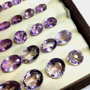 K&ouml;nnte beinhalten: Eine Sammlung oval geschliffener Amethyst-Edelsteine in einem cremefarbenen Tablett. Die Edelsteine zeigen eine Reihe von lilafarbenen Farbt&ouml;nen, von hellem Lavendel bis zu tiefem Violett, mit Facetten, die das Licht einfangen. Das Tablett hat einen dunkelbraunen Rand.