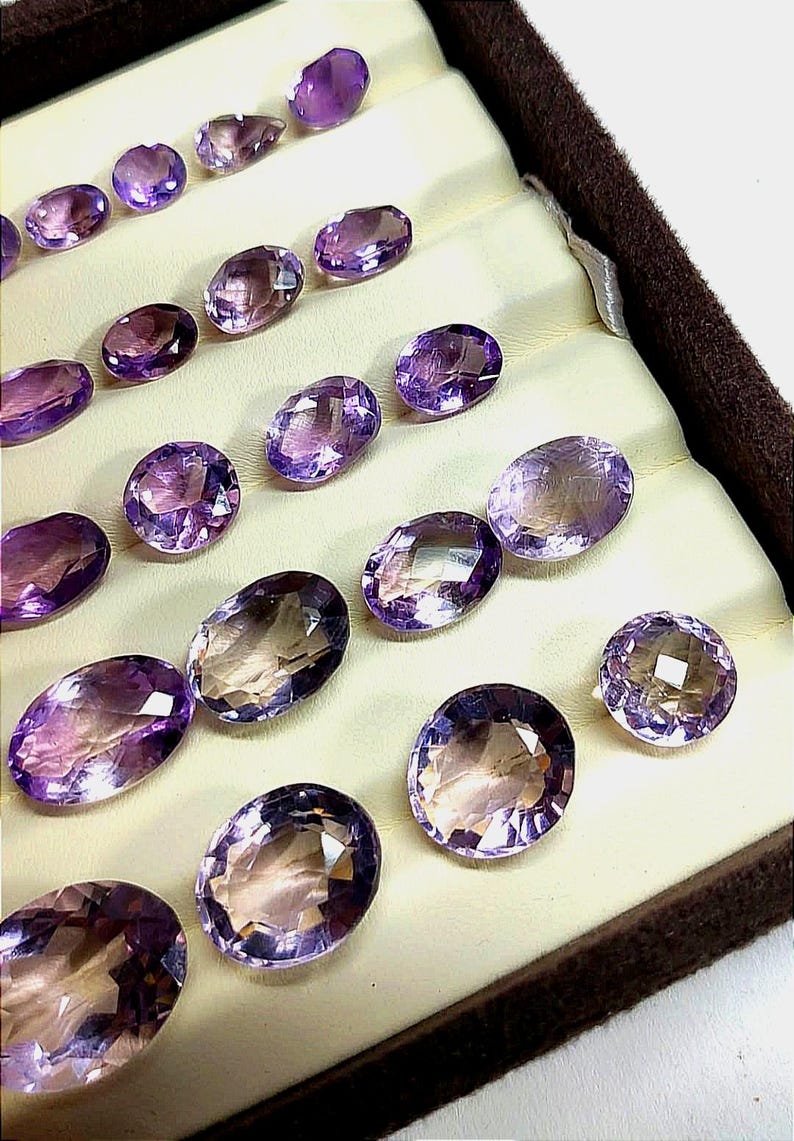 K&ouml;nnte beinhalten: Eine Sammlung oval geschliffener Amethyst-Edelsteine in einem cremefarbenen Tablett. Die Edelsteine zeigen eine Reihe von lilafarbenen Farbt&ouml;nen, von hellem Lavendel bis zu tiefem Violett, mit Facetten, die das Licht einfangen. Das Tablett hat einen dunkelbraunen Rand.