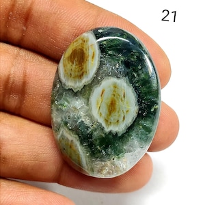 May include: An oval-shaped polished gemstone featuring a deep green translucent base with distinct white and golden-brown circular inclusions. The stone has a smooth, reflective surface and is marked with the number 21 in the upper right corner.