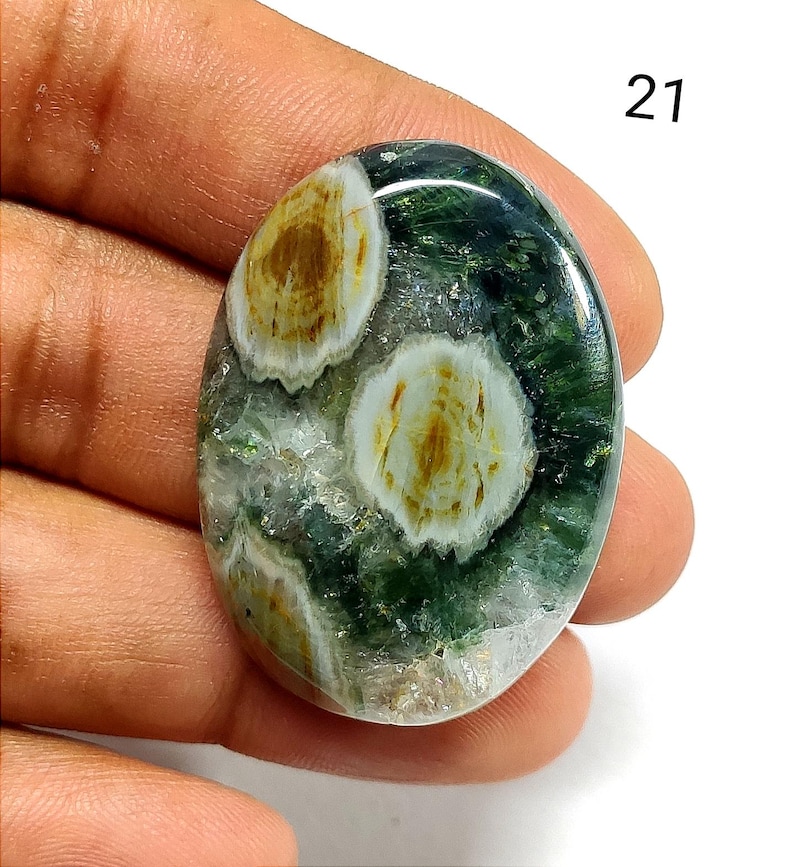May include: An oval-shaped polished gemstone featuring a deep green translucent base with distinct white and golden-brown circular inclusions. The stone has a smooth, reflective surface and is marked with the number 21 in the upper right corner.