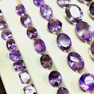 K&ouml;nnte beinhalten: Eine Auswahl an polierten Amethyst-Edelsteinen in verschiedenen Formen, darunter ovale und quadratische Schliffe. Die Edelsteine sind auf einer cremefarbenen Oberfl&auml;che angeordnet und zeigen ihre tiefvioletten Farbt&ouml;ne und reflektierenden Facetten. Eine Sammlung von Schmuckkomponenten.