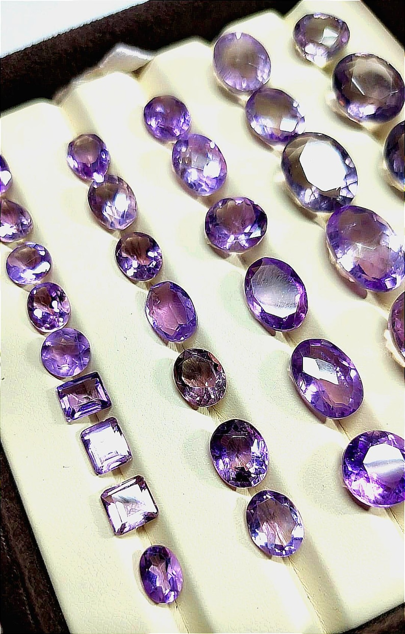 K&ouml;nnte beinhalten: Eine Auswahl an polierten Amethyst-Edelsteinen in verschiedenen Formen, darunter ovale und quadratische Schliffe. Die Edelsteine sind auf einer cremefarbenen Oberfl&auml;che angeordnet und zeigen ihre tiefvioletten Farbt&ouml;ne und reflektierenden Facetten. Eine Sammlung von Schmuckkomponenten.