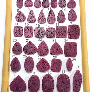 Könnte beinhalten: Eine Sammlung von vierunddreißig unregelmäßig geformten, tiefmagentafarbenen Edelsteinen, jeweils mit poröser Textur. Die Edelsteine sind auf einer weißen Oberfläche in einer Holzrahmen-Anzeige angeordnet.