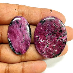 May include: Two oval-shaped ruby zoisite cabochons with a polished finish. The stones feature a vibrant magenta and deep pink base color with scattered black and dark green mineral inclusions. The stones are labeled with the numbers 1 and 2.