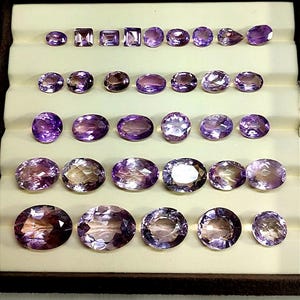 K&ouml;nnte beinhalten: Eine Sammlung von Amethyst-Edelsteinen in verschiedenen Formen und Gr&ouml;&szlig;en, darunter ovale, quadratische und runde Schliffe. Die Edelsteine reichen in der Farbe von hell- bis tiefviolett und werden in einer braunen Schale mit cremefarbenem Innenraum pr&auml;sentiert.