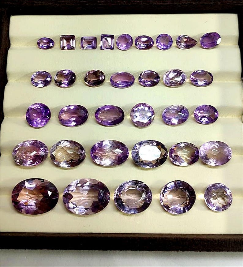 K&ouml;nnte beinhalten: Eine Sammlung von Amethyst-Edelsteinen in verschiedenen Formen und Gr&ouml;&szlig;en, darunter ovale, quadratische und runde Schliffe. Die Edelsteine reichen in der Farbe von hell- bis tiefviolett und werden in einer braunen Schale mit cremefarbenem Innenraum pr&auml;sentiert.
