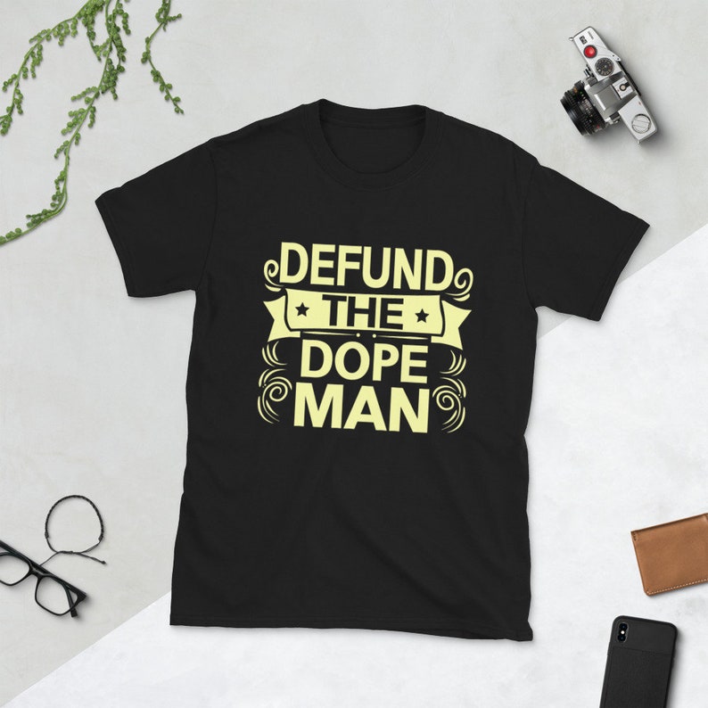 Peut inclure: T-shirt noir avec un graphisme jaune qui dit "Defund the Dope Man" dans une police de caract&egrave;res stylis&eacute;e.