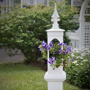 May include: White planter with a decorative finial on top, holding a pot of purple flowers. The planter is attached to a white post.
