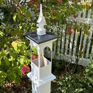 May include: White post with a black top and a white finial. The post has a decorative archway design and is surrounded by green foliage.
