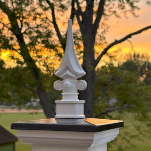 May include: White decorative finial with a pointed top and intricate details. The finial is mounted on a black square base.