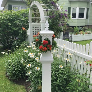 May include: A white picket fence with a white wooden post topped with a decorative planter. The planter is filled with orange and red flowers. The fence is in front of a green lawn and a house with a white trellis covered in purple flowers.