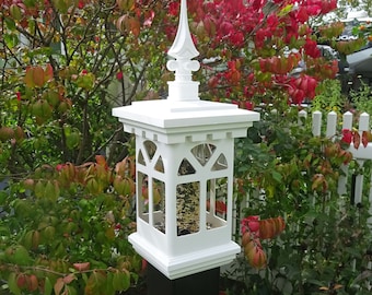 May include: White bird feeder with a decorative finial on top. The feeder is made of wood and has a clear plastic window. The feeder is mounted on a black post. The feeder is surrounded by red and green foliage.