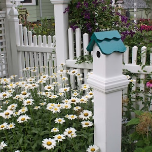 May include: A white birdhouse with a teal roof sits atop a white post in a garden. The birdhouse is surrounded by a white picket fence and a bed of daisies with white petals and yellow centers. Purple flowers and greenery are in the background.