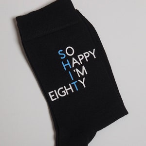 May include: Black socks with white text that reads "So Happy I'm Eighty" in a vertical design. The word "Happy" is in blue.