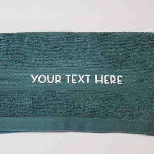 May include: A teal hand towel with a white embroidered border and the text "YOUR TEXT HERE" in white.