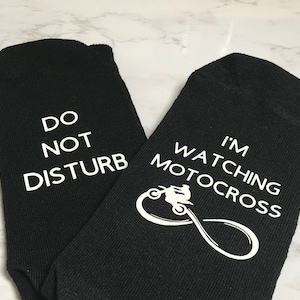 May include: A pair of black socks with white text. One sock says "Do Not Disturb" and the other says "I'm Watching Motocross" with a silhouette of a motocross rider.