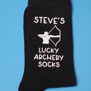 May include: Black socks with white text that reads "STEVE'S LUCKY ARCHERY SOCKS". A white silhouette of an archer with a bow and arrow is printed on the socks.