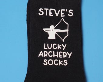 May include: Black socks with white text that reads "STEVE'S LUCKY ARCHERY SOCKS". A white silhouette of an archer with a bow and arrow is printed on the socks.