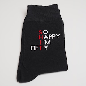 May include: A black sock featuring white text that reads SO HAPPY I'M FIFTY, with the letters S, H, I, T highlighted in red to form a vertical word. The sock has a ribbed cuff at the top and a standard crew length design with a flat, folded appearance.