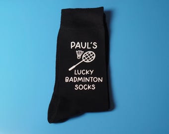 May include: Black socks with the text "PAUL'S LUCKY BADMINTON SOCKS" in white. The design includes a badminton shuttlecock and racket. The socks are on a blue background.