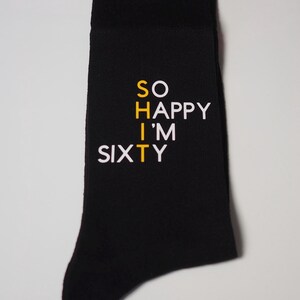 May include: Black socks with white text that reads "SO HAPPY I'M SIXTY" with the "T" in "SIXTY" in yellow.