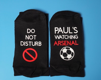 May include: A pair of black socks with white text. One sock says "Do Not Disturb" with a red circle and diagonal line through it. The other sock says "Paul's Watching Arsenal" with a white soccer ball.