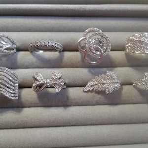 May include: Eight silver rings with clear cubic zirconia stones. The rings feature different designs, including a wave, a bow, a leaf, and a geometric pattern.