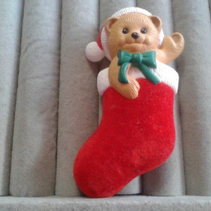 May include: A red felt Christmas stocking with a brown teddy bear wearing a white Santa hat and a green bow.