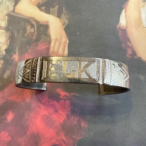 May include: Silver-toned cuff bracelet with the word "ELK" engraved in the center. The bracelet has geometric patterns on the sides. The background features a painting with muted colors.