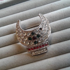 Puede incluir: Un broche de plata en forma de media luna con diamantes de imitación transparentes, rojos y verdes.