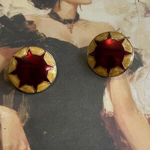 Puede incluir: Un par de pendientes de estrella roja y dorada. Los pendientes son redondos y tienen un borde dorado con un centro rojo. El centro de los pendientes es un diseño de estrella con detalles dorados.