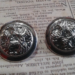 Könnte beinhalten: Ein Paar silberfarbene Ohrringe mit einem strukturierten, floralen Design. Die Ohrringe sind rund und haben einen erhöhten Rand.