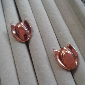 May include: Two copper-colored tulip-shaped earrings. The earrings have a polished, reflective surface and are set against a gray, textured background. The tulip design features curved petals and a central bud, creating a floral theme.