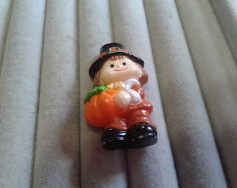 May include: A small figurine of a pilgrim holding a pumpkin. The pilgrim is wearing a brown and black hat and a brown and orange outfit. The pumpkin is orange with a green stem.