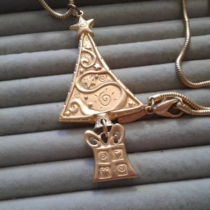Könnte beinhalten: Eine goldfarbene Halskette mit einem detaillierten Weihnachtsbaum-Anhänger und einem kleinen Geschenkbox-Anhänger. Die Kette ist im Schlangenketten-Stil.